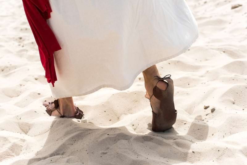 Jesus enters a town with a cropped image showing base of white robes and red shawl and feet in sandals, walking on sand.