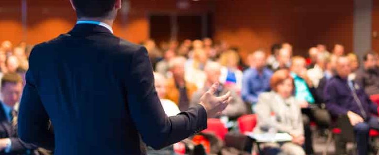 Events photo of back of man in a black suit standing in front of a seated audience in a large conference room.